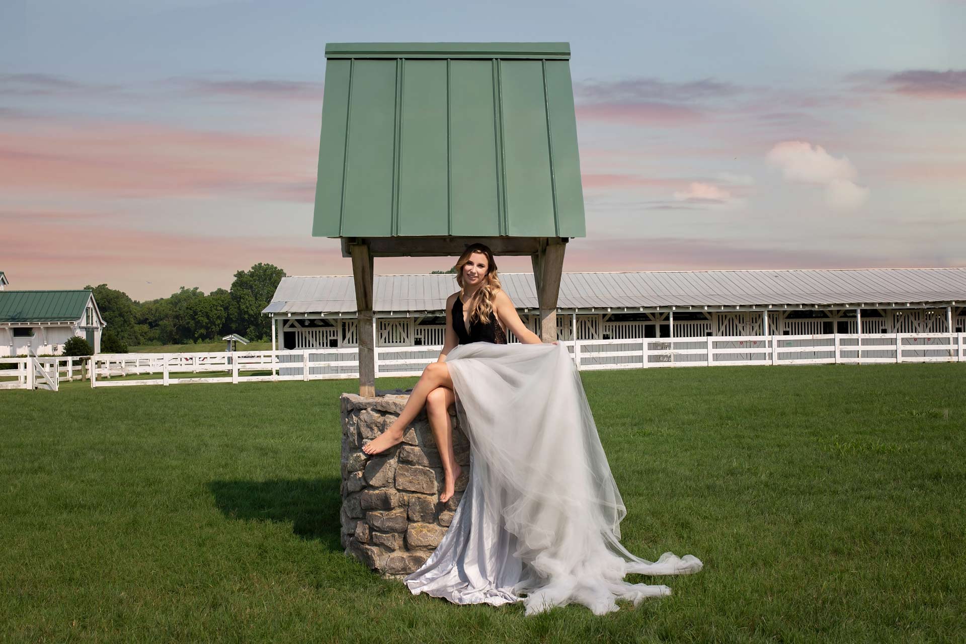 High school senior wearing a long tulle skirt and sitting on top of a wishing well during a professional senior photography session at Moments By T, Franklin Tennessee