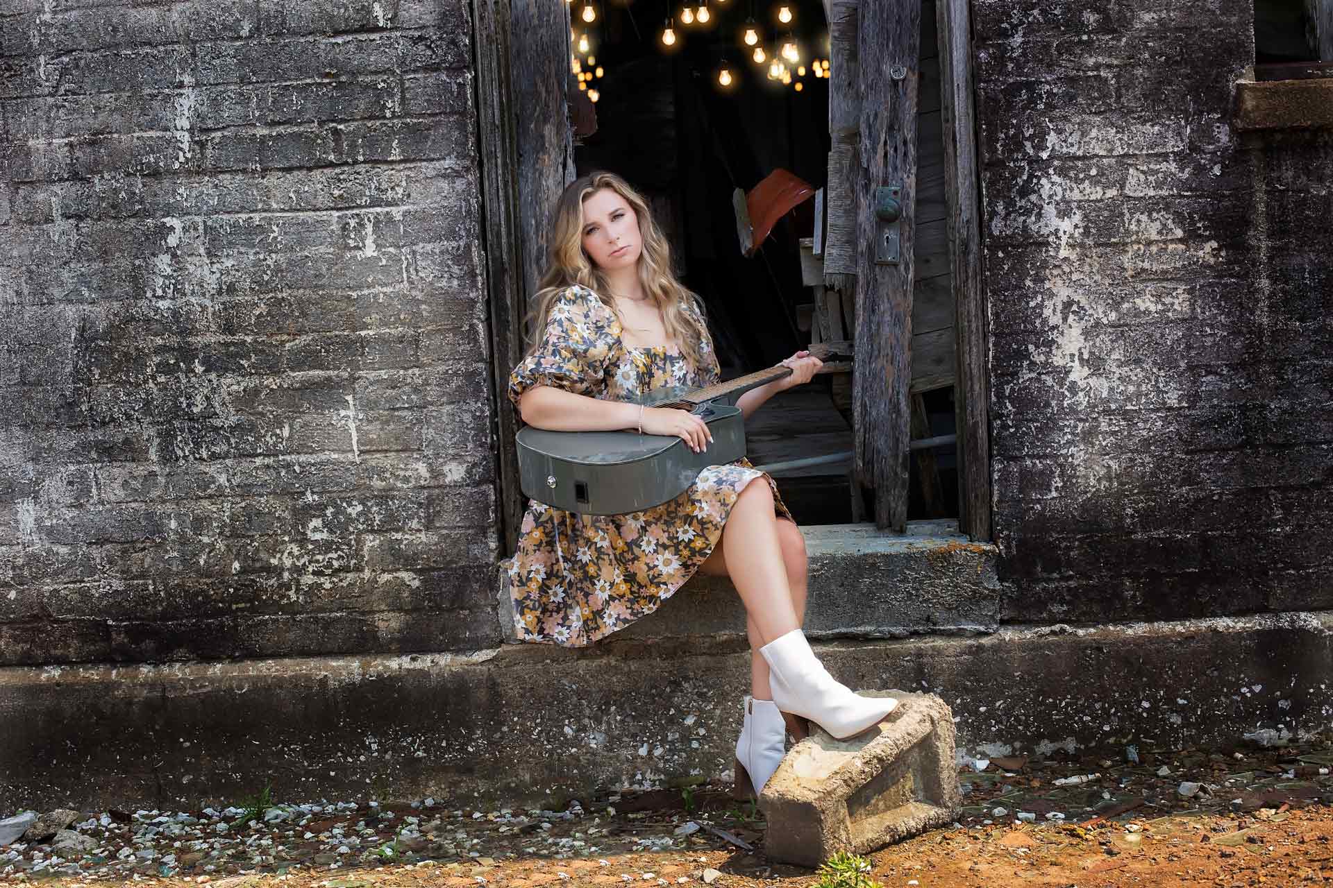 blonde high school senior sitting in a brick barn doorway with her guitar during a professional senior photography session at Moments By T, Franklin Tennessee