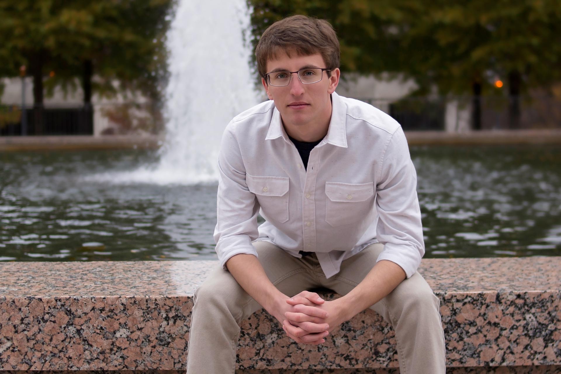 High school senior boy sitting in front of a fountain with his hands folded on his lap during a professional senior photography session at Moments By T, Franklin Tennessee