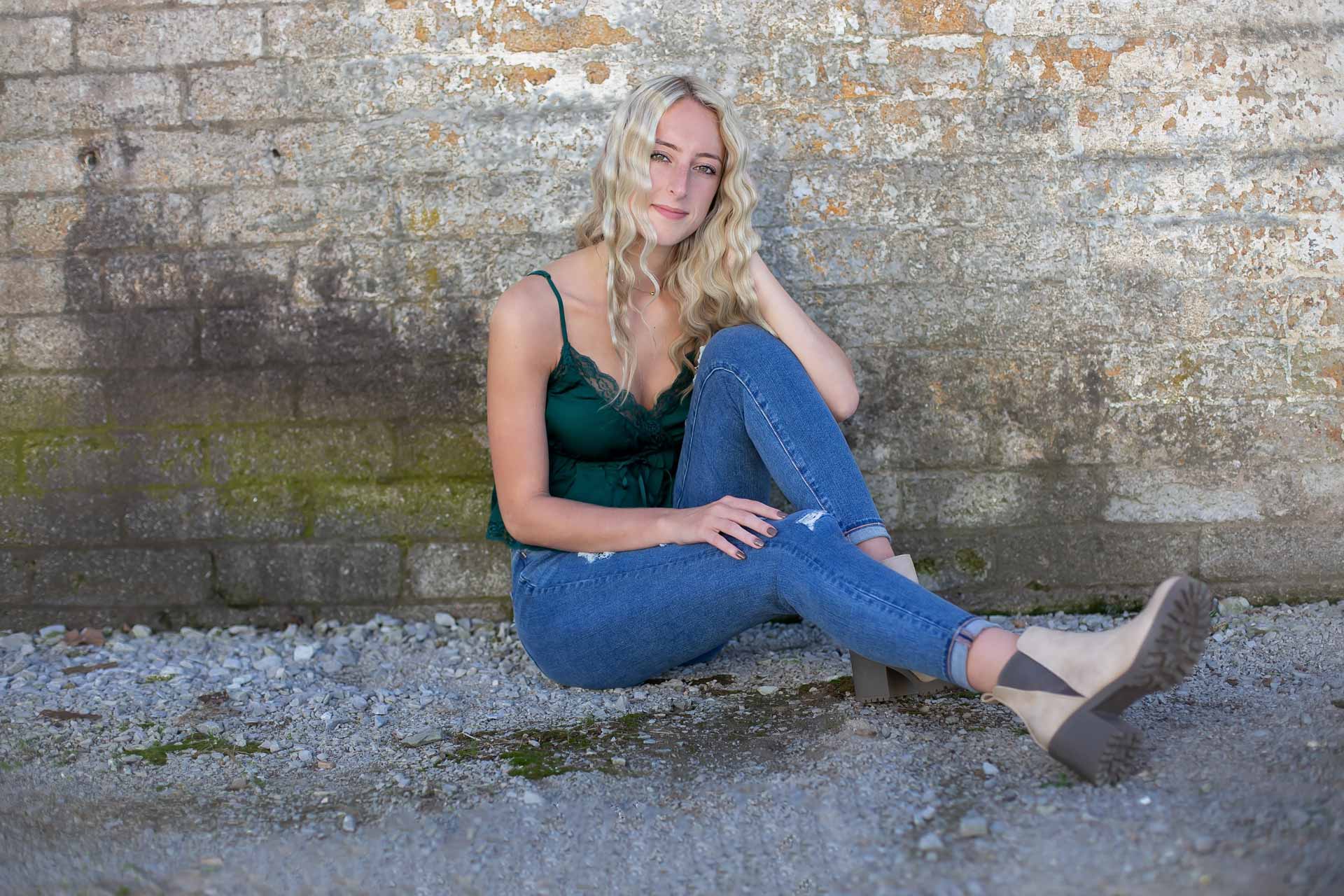 blonde senior girl sitting against a white dirty brick wall during a professional senior photography session at Moments By T, Franklin Tennessee