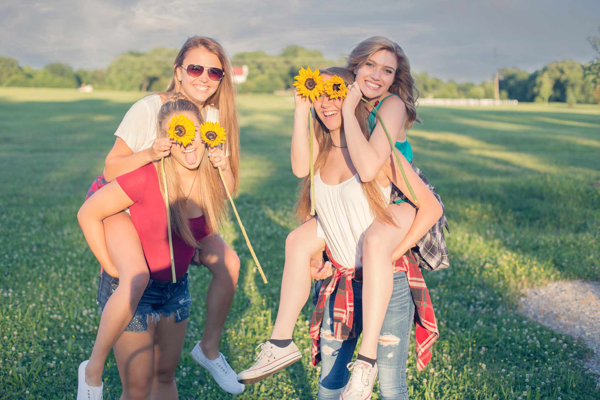 Group of four teenage girls playing piggy back rides while holding sunflowers on their eyes during a professional senior photography session at Moments By T, Franklin Tennessee