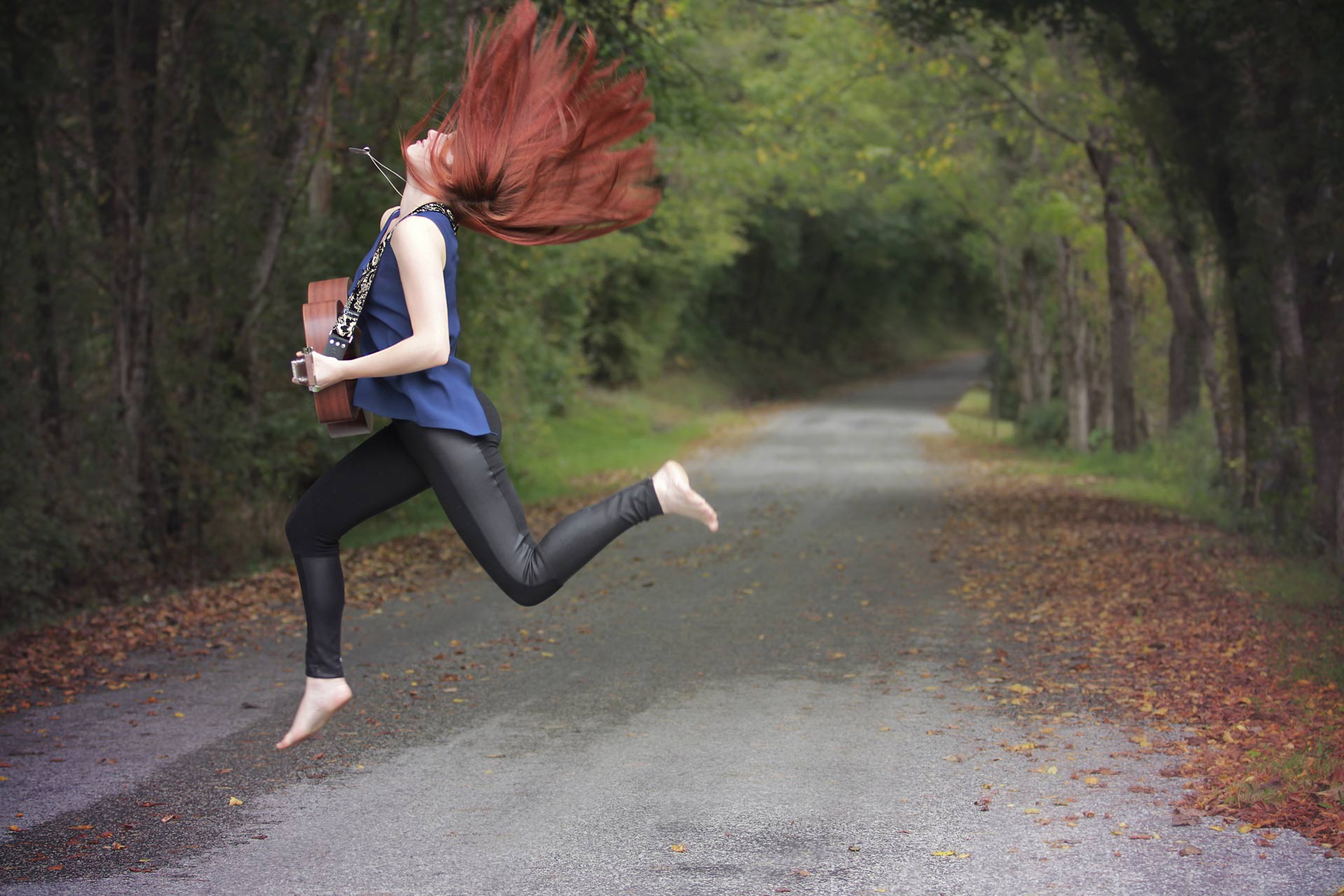 Redhead high school senior jumping in the air holding her guitar in the middle of a road surrounded by trees during professional senior photography session at Moments By T, Franklin Tennessee