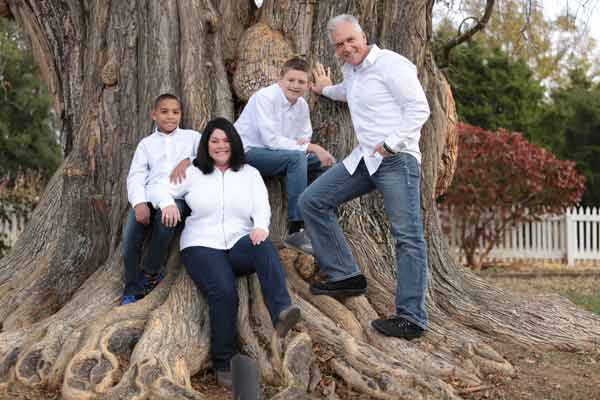 Family of four standing in front of an old tree wearing white white shirts and jeans during their family photoshoot