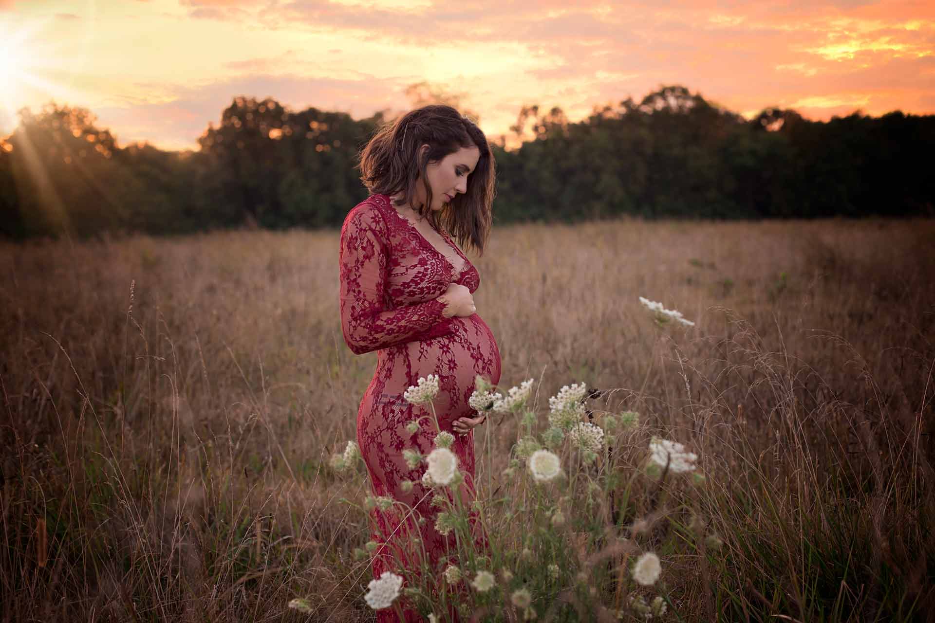  pregant woman wearing a red see through robe standing in a field of flowers during sunset